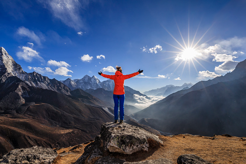 Young woman, wearing red jacket, lifts her arms in victory. She is standing on the top of a mountain and watching sunset over Himalayas .Mount Everest National Park. This is the highest national park in the world, with the entire park located above 3,000 m ( 9,700 ft).