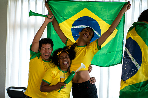 soccer fans, men and women from brazil, are at the house of one of them dressed in flags team jerseys, watching a soccer game while hugging each other and celebrating a great gollll