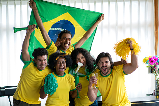 soccer fans, men and women from brazil, are at the house of one of them dressed in flags team jerseys, watching a soccer game while hugging each other and celebrating a great gollll