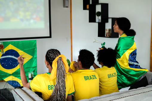soccer fans, men and women from brazil, are in the house of one of them in the living room dressed in flag team jerseys, watching a soccer game with great care and concentration