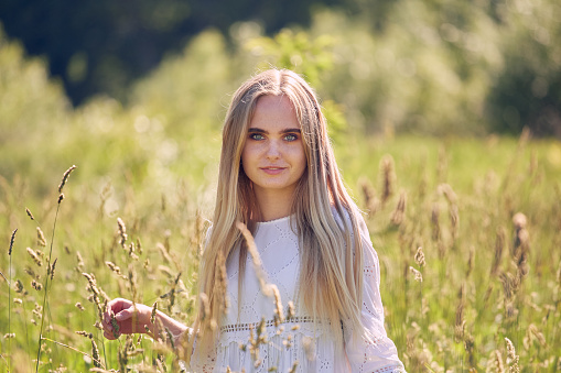 happy young blond woman in white dress at sunny meadow looking at camera smiling