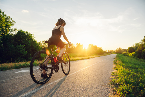 Rear view of a young woman cyclist riding road bicycle on a free road in the forest at a sunny day. Healthy lifestyle concept. Sunset in the background. Copy space.