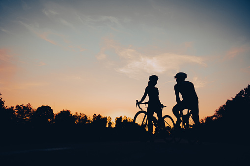 Silhouettes of a professional riders cycling on racing bikes on the road in nature during sunset. Active lifestyle and sport concept. Sporty couple of cyclist. Copy space.