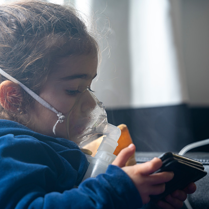 Close up photo of 3 years old girl sitting in living room and using nebulizer for allergy treatment. Shot indoor with a full frame mirrorless camera.