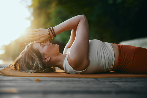 A mindful woman is lying on her back and holding hands in a namaste position while practicing breathing on the dock near the river in nature.