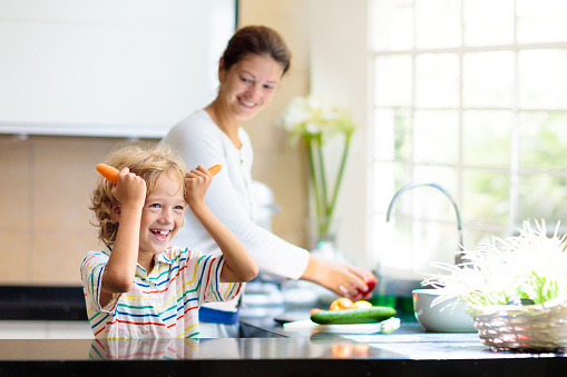 Mother and child make dinner in white kitchen. Family at home. Mom and little boy cut fresh vegetables for healthy lunch. Parents and kids bonding. Young woman and her kid cook meal.
