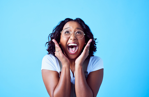 Excited, happy and surprised young female is amazed while looking up. Delight on a black or African woman's face against a bright blue background. Trendy and stylish lady celebrating being a winner