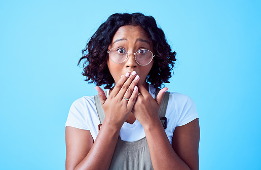 Surprised, shocked and excited woman looking amazed and showing funny expression standing inside. Face of curious, african female hearing interesting news with hands covering mouth, isolated on blue