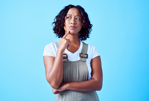Confused, thinking and daydreaming woman against a blue background with copy space. Young black female looking bored, planning something fun, lost in thought. African American lady needs a change