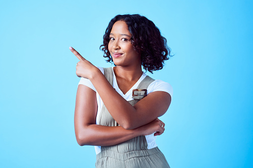 Woman pointing her finger at corner on her right showing copy space for ad space against a bright blue background  in studio. Friendly smiling lady points to free advertising space to market a sale