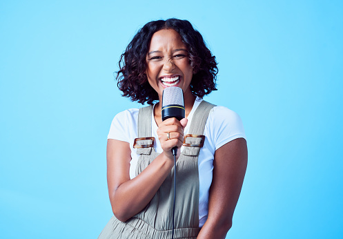 Karaoke, podcast and singing woman with a microphone against a bright blue background. Portrait of a young playful, excited and happy woman holding a mic. Delighted lady enjoying a broadcast
