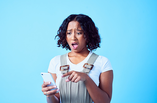 OMG, shocked and speechless woman pointing to her phone in disbelief after reading gossip or news online. Young female gasping in awe at a text message, meme or internet post on her social media app
