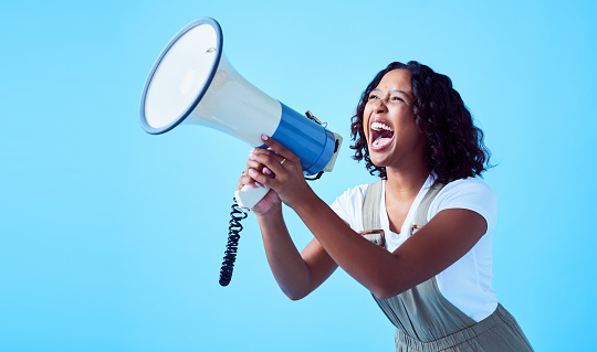 Funny, excited and crazy female screaming on a megaphone against a bright blue background. A young loud and happy woman announcing on a loudspeaker Cheerful lady making an announcement