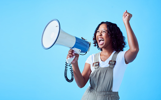Happy, excited woman cheering on a megaphone, making an announcement. African American female celebrating a win or victory, smiling and shouting against a blue, copyspace background.