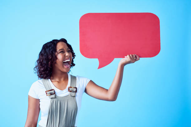 istock Woman screaming into a speech bubble with emotion on her face and against a blue background with copy space. Lady shouting and yelling into a social media board to chat a message inside a studio 1414324484