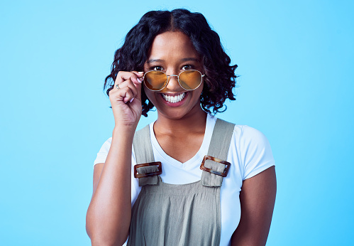 Happy, trendy and stylish woman wearing cool sunglasses, smiling in an isolated studio portrait against a blue background. Confident, friendly and excited young lady smiles showing her happiness
