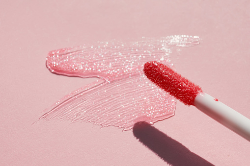Smears of shining lip gloss and lip gloss brush on pink background, hard shadows.