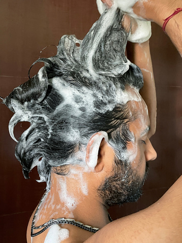 Stock photo showing side view of Indian man making shampoo soap sud quiff whilst washing his hair in a wet room shower.