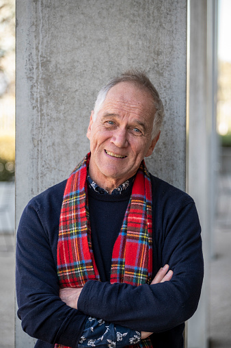 Smiling older man in tartan scarf looks at camera. He waits with folded arms.