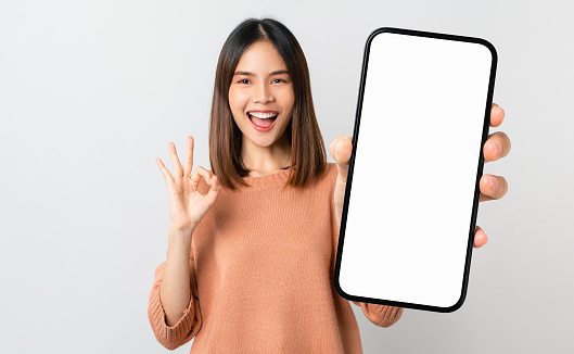 Hermosa mujer asiática sosteniendo una maqueta de teléfono inteligente de pantalla en blanco y muestra un signo de ok sobre fondo gris photo