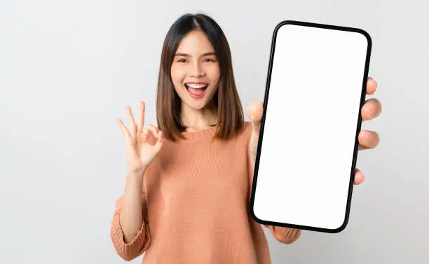 Photo of Beautiful Asian woman holding smartphone mockup of blank screen and shows ok sign on grey background