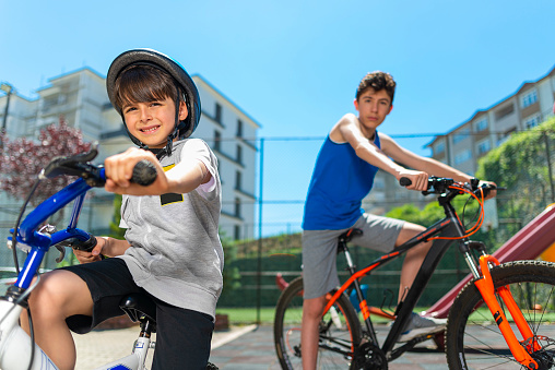 His primary school-age brother and his high-school-age brother, who wear safety helmets, are riding bicycles. Happy children in colorful clothes riding bikes in front of playground in the city. Outdoor cycling is a healthy activity for kids.