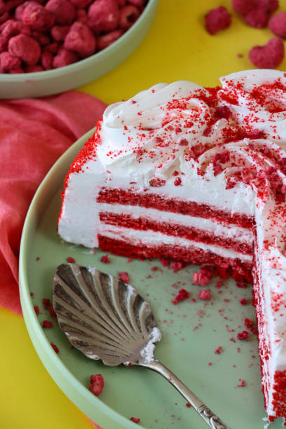 Stock photo showing close-up, elevated view of pastel green plate containing a sliced, red velvet cake displaying layers of red sponge and white butter cream. This dessert is decorated with piped icing swirls and dusted with dehydrated raspberry powder. imagen de primer plano pastel de terciopelo rojo en rodajas sobre plato verde pastel, pastel de capas con crema de mantequilla, decorado con remolinos de glaseado entubado, polvo de frambuesa deshidratado, servidor de pasteles, tazón de frambuesas deshidr