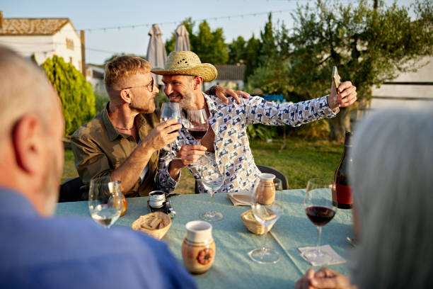 Mature gay couple taking selfie at outdoor wine tasting Over the shoulder view of relaxed men sitting at table, holding glasses of red wine, and smiling face to face as they capture a memory. tourist couple candid travel stock pictures, royalty-free photos & images