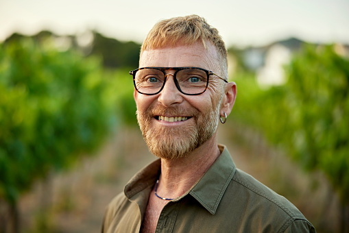 Head and shoulders view of early 50s man with strawberry blond hair wearing eyeglasses, open collar shirt, and smiling at camera between grapevines.