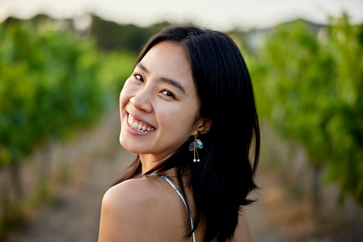 Close view of woman with long black hair wearing summer dress and pausing from walking between grapevines to look over shoulder and smile at camera.
