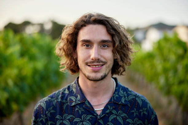 Portrait of early 20s man standing in vineyard Head and shoulders view of man with medium-length brown hair, mustache and beard, wearing open collar shirt and smiling at camera with grapevines in background. mid length hair stock pictures, royalty-free photos & images