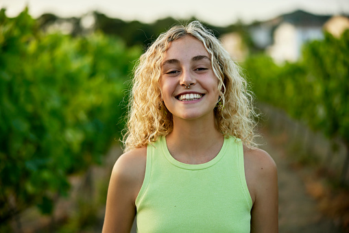 Head and shoulders view of wine lover with long curly blond hair wearing chartreuse tank top and grinning at camera with grapevines in background.
