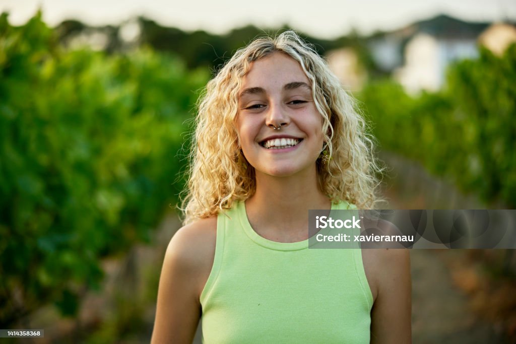 Portrait of woman in late teens standing in vineyard Head and shoulders view of wine lover with long curly blond hair wearing chartreuse tank top and grinning at camera with grapevines in background. Teenager Stock Photo