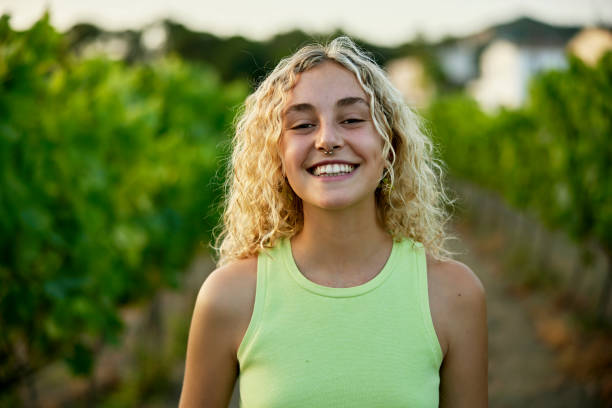 Portrait of woman in late teens standing in vineyard Head and shoulders view of wine lover with long curly blond hair wearing chartreuse tank top and grinning at camera with grapevines in background. mediterranean woman portrait stock pictures, royalty-free photos & images