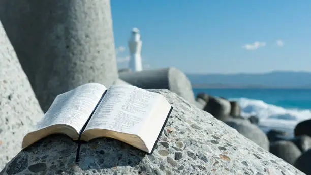 Holy Bible open to Psalm 119 on top of a breakwater. Beautiful blurred background with white lighthouse and sea with waves.