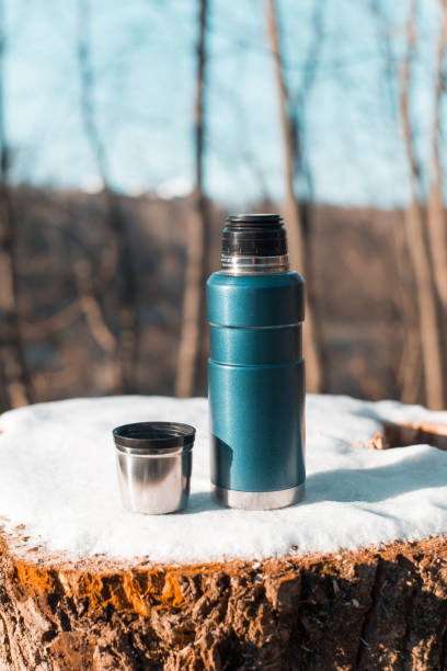 istock Thermos and a mug with a hot drink standing on a stump. Winter forest warming drink in a cup. Vertical photo 1414371417