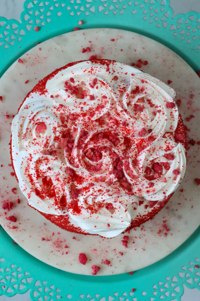 Stock photo showing close-up, elevated view of white, cake cutting board on patterned turquoise blue cake stand containing a whole, red velvet cake covered in butter cream and decorated with piped icing swirls dusted with dehydrated raspberry powder. imagen en primer plano de un pastel de terciopelo rojo entero cubierto con crema de mantequilla, decorado con remolinos de glaseado entubado y polvo de frambuesa deshidratado, tablero de pastel blanco en un soporte de pastel azul turquesa estampado, vista