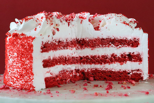 Stock photo showing close-up view of white cake cutting board on patterned turquoise blue cake stand containing a cut, red velvet cake covered in butter cream and decorated with piped icing swirls dusted with dehydrated raspberry powder.