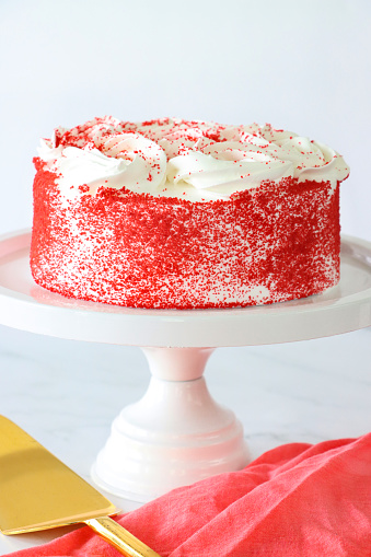 Stock photo showing close-up view of white cake stand containing a whole, red velvet cake covered in butter cream and decorated with piped icing swirls dusted with dehydrated raspberry powder.