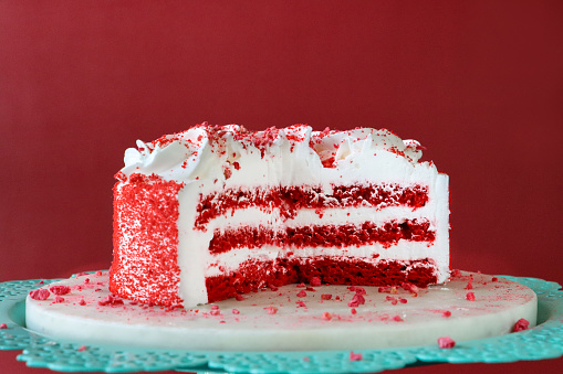 Stock photo showing close-up view of white cake cutting board on patterned turquoise blue cake stand containing a cut, red velvet cake covered in butter cream and decorated with piped icing swirls dusted with dehydrated raspberry powder.