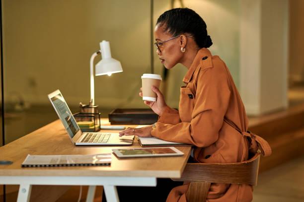 Creative black entrepreneur or freelancer working on a laptop at night, multitasking holding coffee cup. Dedicated designer completing project late, sitting at a desk in home office with lamp light.