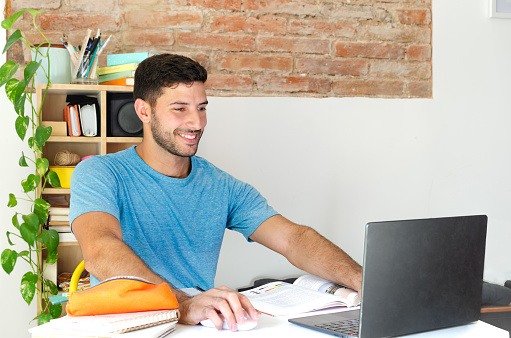 young man smiles while using his computer at home. he is looking for information on the internet on his small desk. back to school concept with copy space. entrepreneur working from home.