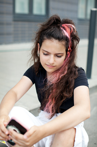 Thoughtful Teenager girl with long dark curly colored hair dyed pink looks into smartphone gadget, chats or video calls
