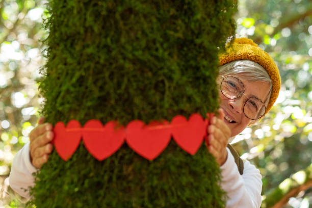 istock Smiling partially hidden elderly woman hugging a moss covered tree trunk in the woods with a wreath of small red hearts - earth day concept. People save the planet from deforestation 1414387356