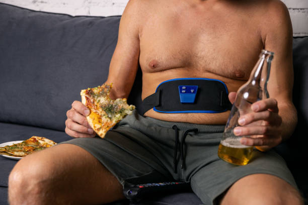 man sitting on couch eat pizza and drink beer while using electronic abdominal slimming belt. unhealthy lifestyle stock photo