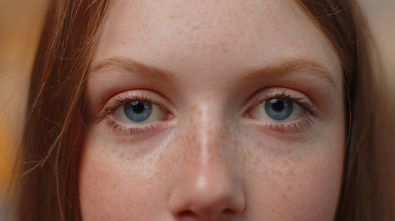 Extreme close-up macro portrait of freckles smiling girl face. Teen beautiful kid's eyes, looking at camera. Blue eyes of redhead female child model. Young positive cute children opening wide her eyes