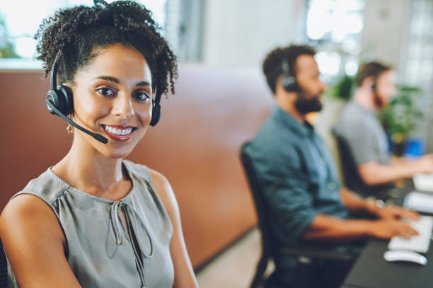 African American female sales team leader or customer service agent working in a call center, talking to a client with a headset. Happy business woman office with diverse colleagues in background. stock photo