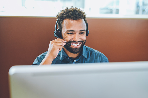 Call center agent talking, wearing a headset and helping people online with a desktop computer in office at work. Young male customer service worker, operator and helpline employee giving support