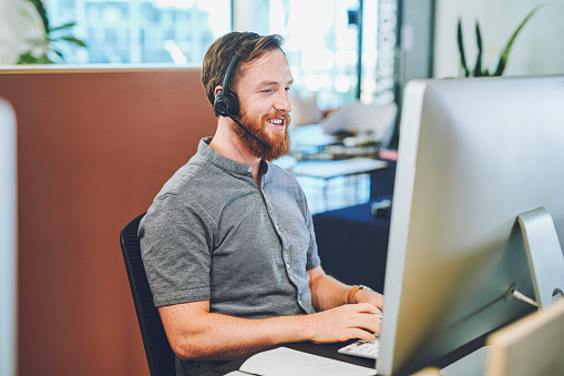 Male call center agent on a laptop working, happy and smiling while talking to a client on a headset in a modern office. A young professional online and phone hotline service operator sitting relaxed
