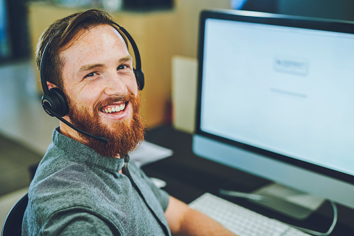 Young man with headphones, at computer, working in tech support or customer care. Happy, smiling male professional call center agent in office. IT technician, telemarketer or online gamer on laptop.